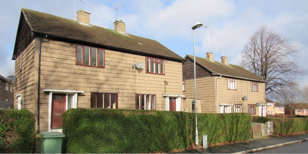 A suburban street with houses that have light brown walls and dark brown roofs, behind green leafy hedges