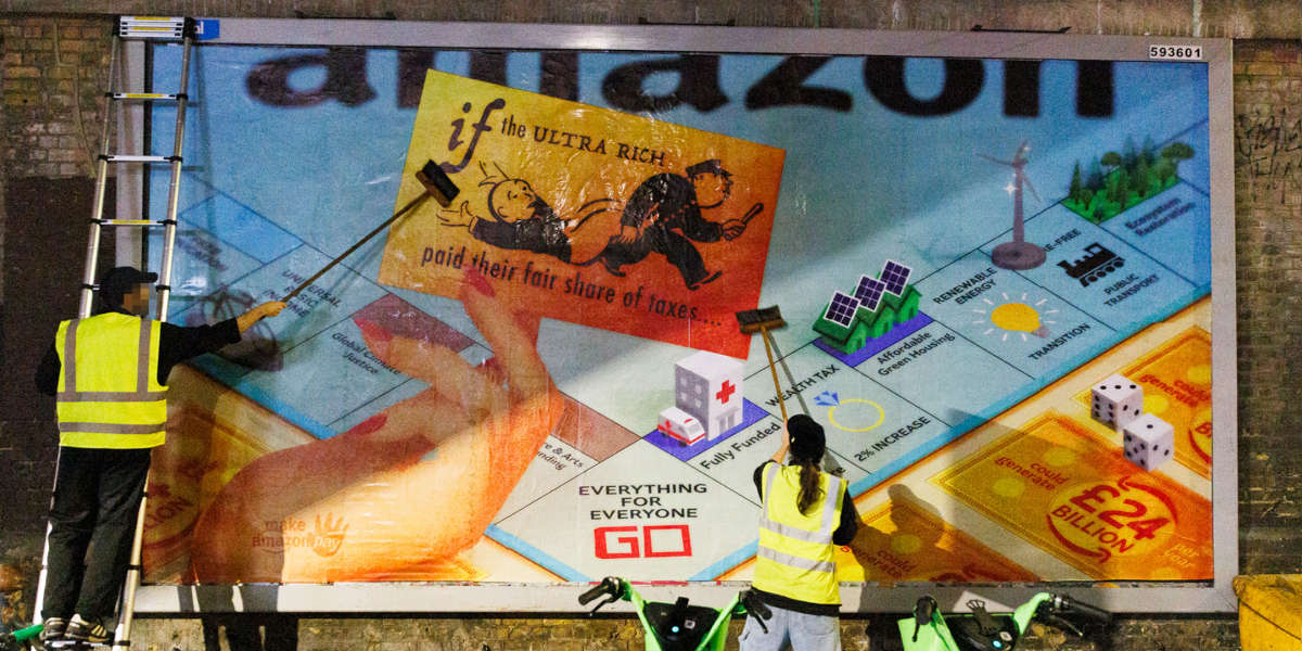 Two people in high-vis yellow vests are pasting up a billboard, which shows a Monopoly board subverted to critique Amazon