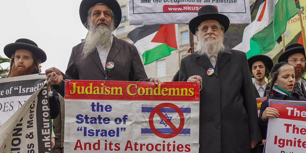A group of Orthodox Jewish men at a protest, with Palestinian flags in the background, holding a placard reading 'Judaism condemns the State of "Israel" and its attrocities'.