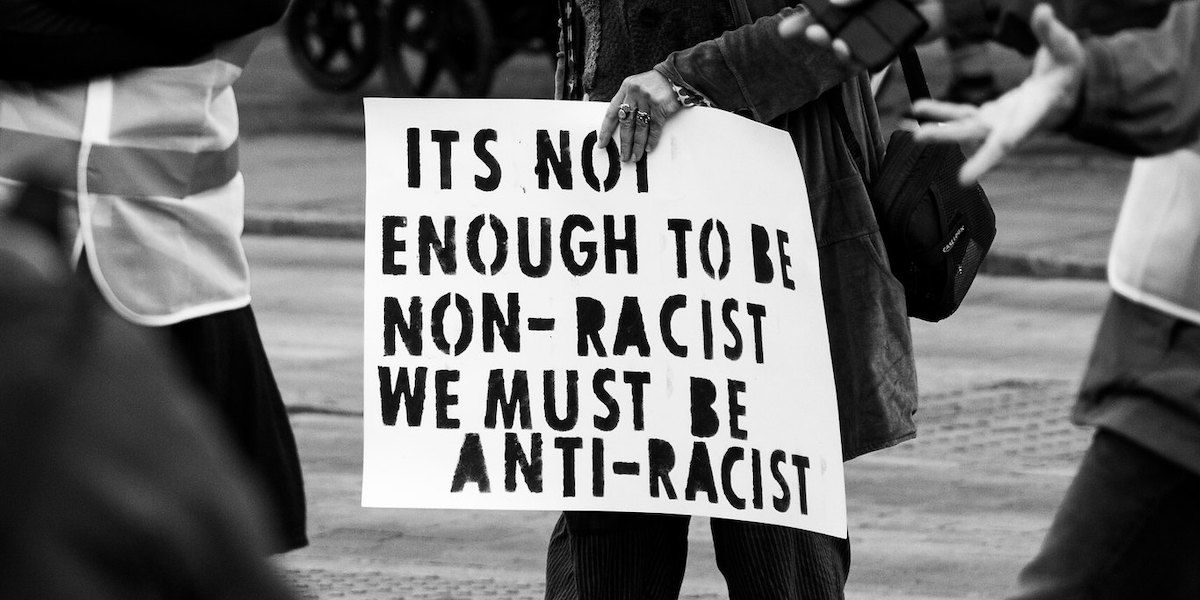 A black and white photo of a person holding a sign that reads: It's not enough to be non-racist we must be anti-racist"