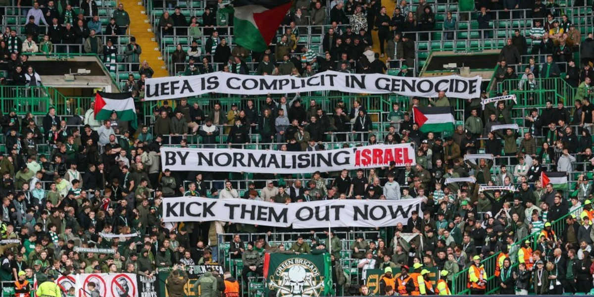 In a football stadium crowd, three strips of banner read: UEFA is complicit in genocide by normalising Israel kick them out now