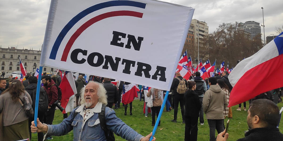 A rally against the proposed Chilean constitution, with Chilean flags in the background and a man holding a large sign reading 'EN CONTRA' (against) in the foreground