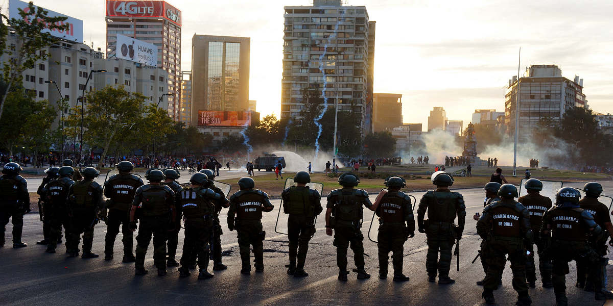 A line of riot police facing away from the camera, protestors and tear gas ahead of them