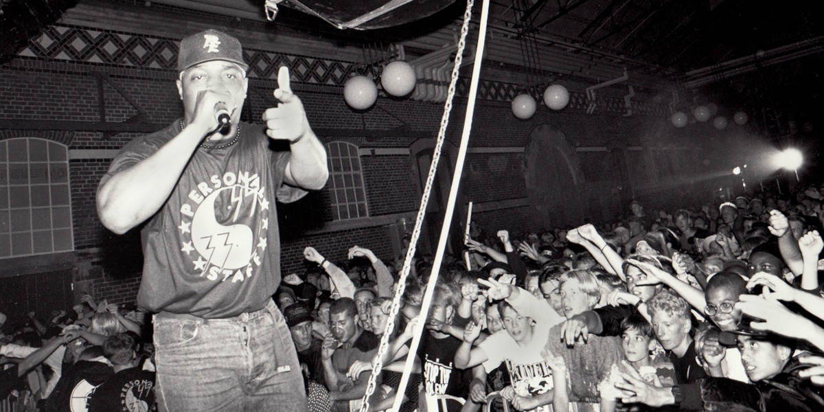 A black and white photo of rapper Chuck D performing on stage, facing away from the crowd and pointing at the camera with a microphone in his other hand