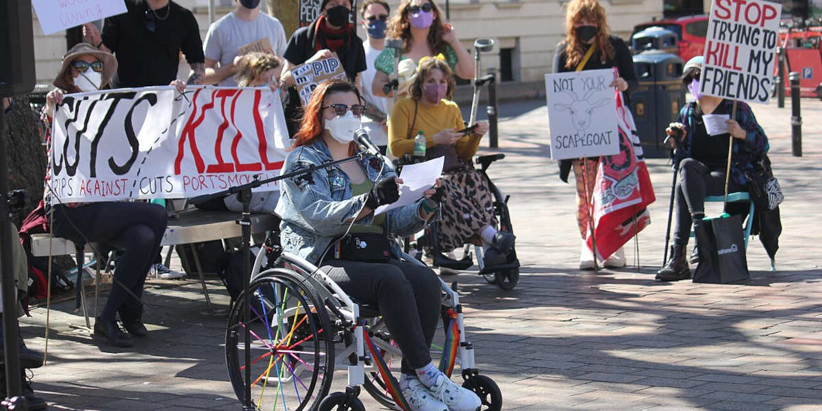A woman wearing a mask in a wheelchair speaking into a microphone, while other protestors holding signs and placards stand behind her