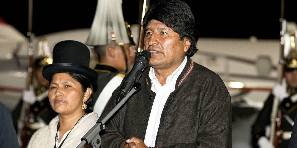 Former Bolivian President Evo Morales stood at a microphone, wearing a white shirt and black jacket. A woman in traditional Bolivian dress stands to his right