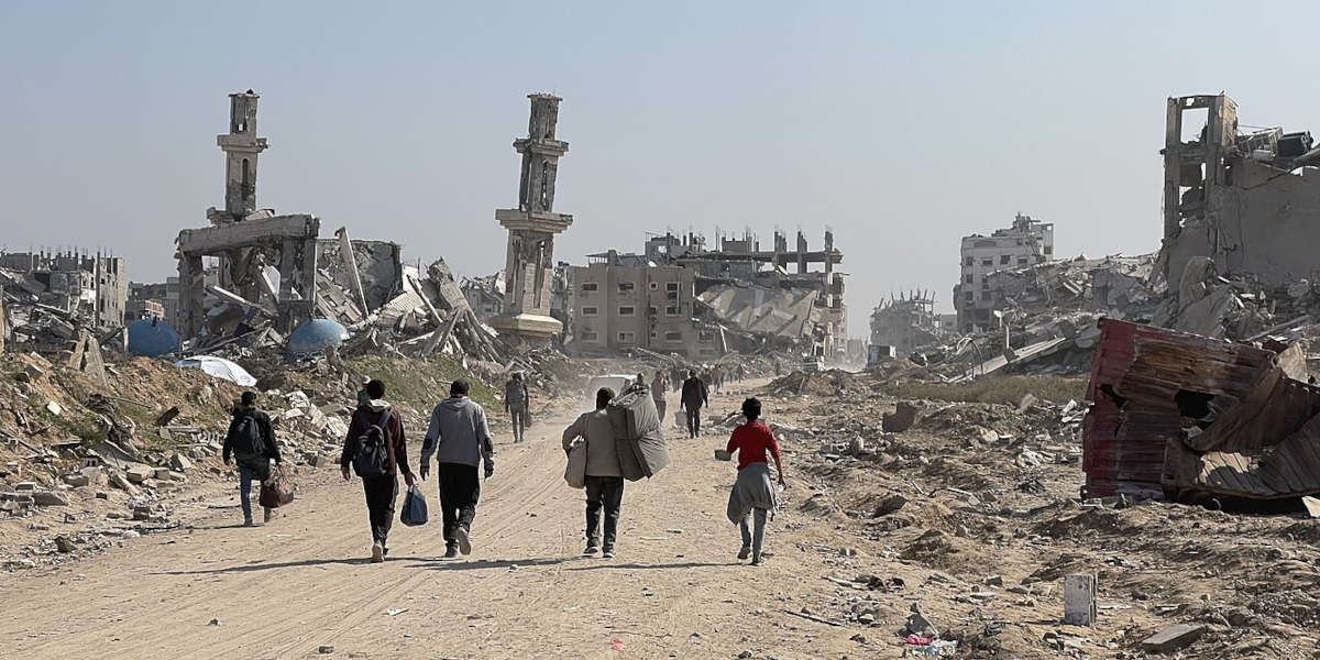 Displaced Gazans walking with their backs turned to the camera and carrying their possessions surrounded by rubble and destroyed buildings on either side of the road they are walking on