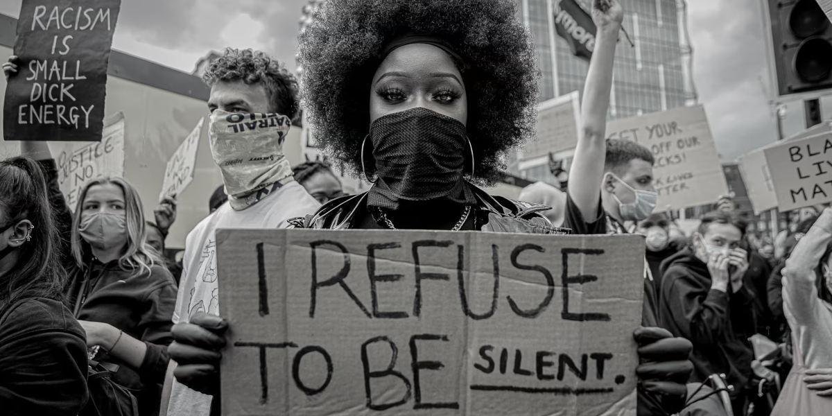 A black and white photo of a woman wearing a mask holding a sign reading 'I refuse to be silent' surrounded by other protestors