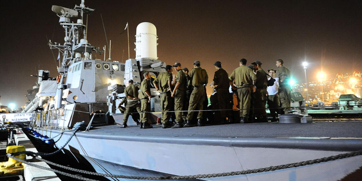 A group of soldiers gathered together on the deck of a ship at night, illuminated by bright lights in the background