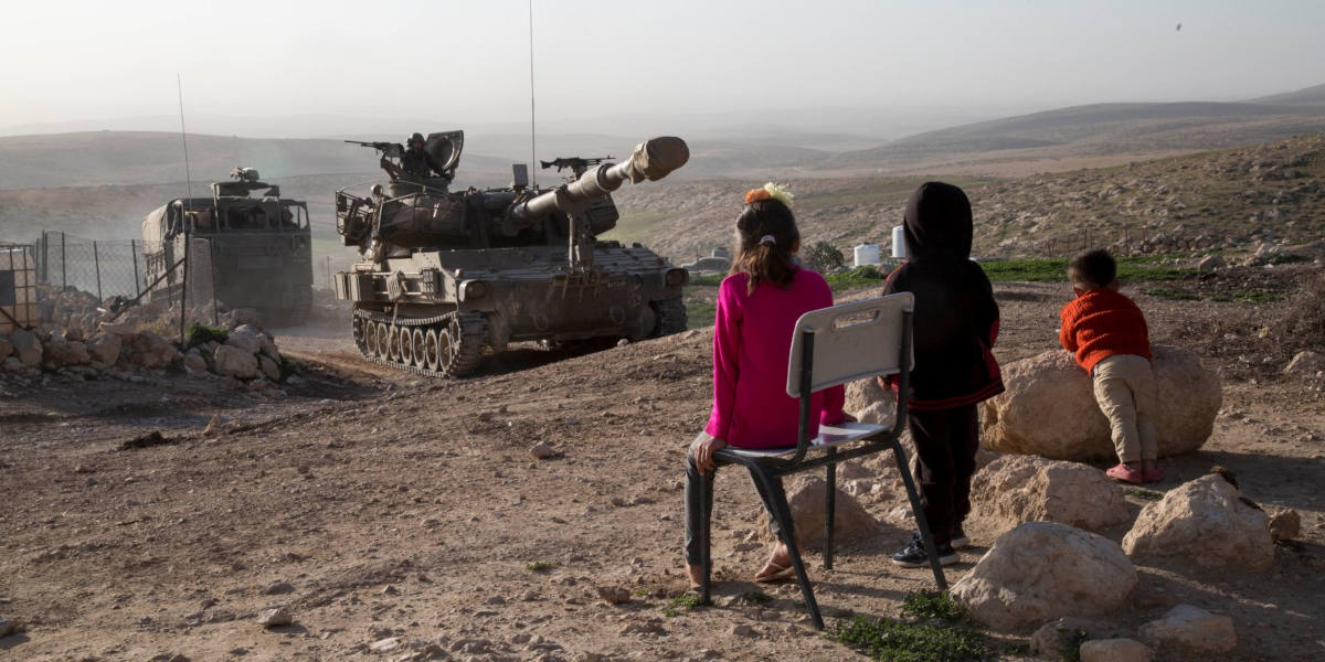 Three Palestinian children with their backs to the camera watching Israeli military vehicles attempting to intimidate them in the West Bank