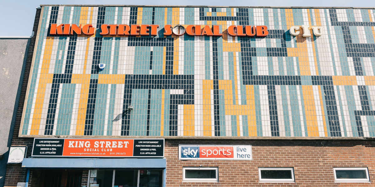 The facade of a building shows 1970s-style tiles with KING STREET SOCIAL CLUB in large letters above. Below a banner for Sky Sports and another King Street sign sit above a brick wall entrance