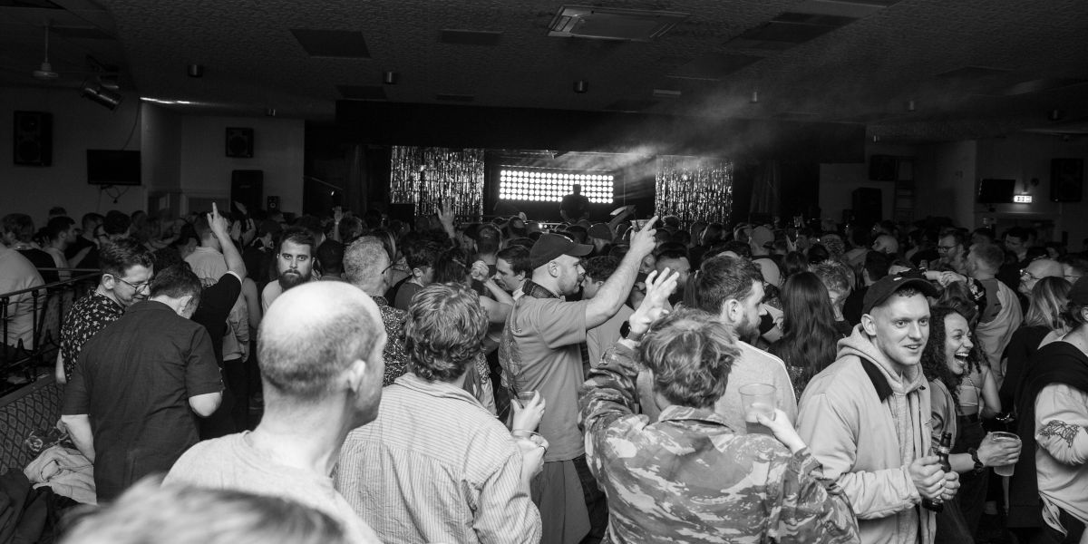 A black and white photo shows a large crowd of sweating revellers with a stage and DJ booth in the distance