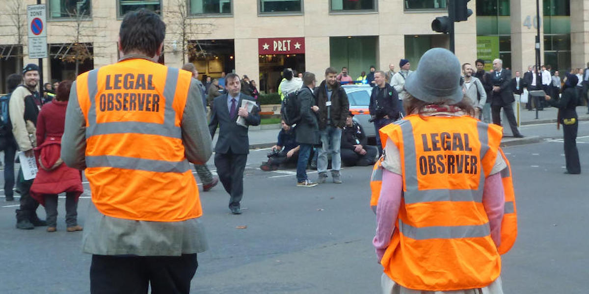 Two people with their backs turned to the camera wearing high-vis orange vests with 'legal observer' written on them