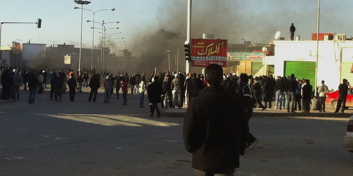 A man with his back to the camera in the foreground, looking towards a crowd of people, whilst a plume of smoke rises further behind them