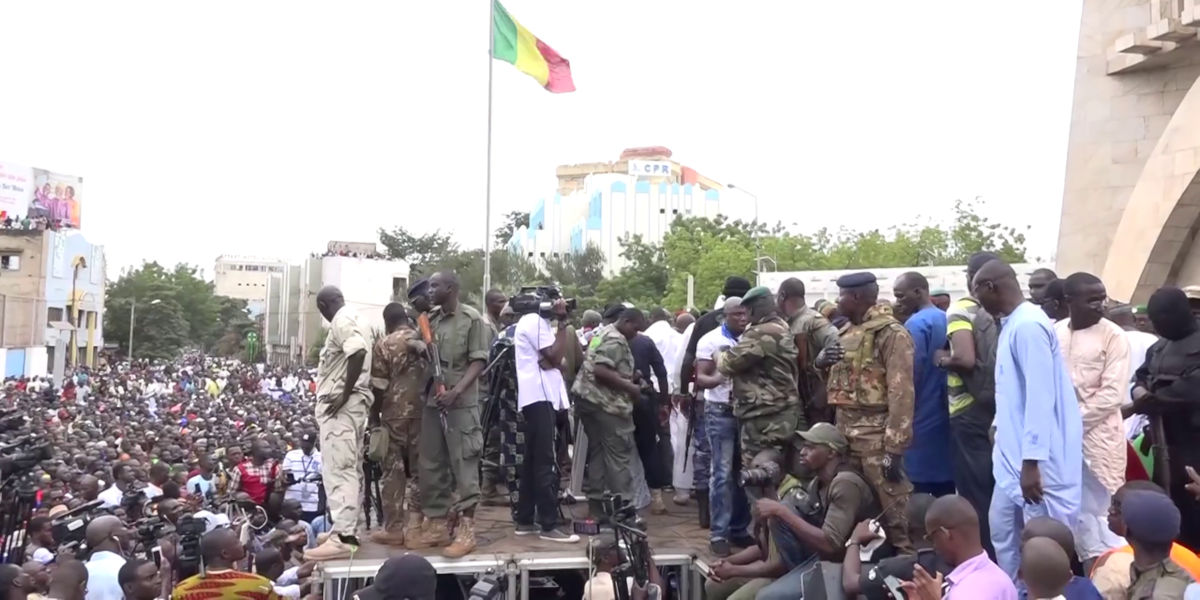 A crowd of people, some dressed in military fatigues, at a protest. The Malian flag can be seen flying on a pole in the centre of the image