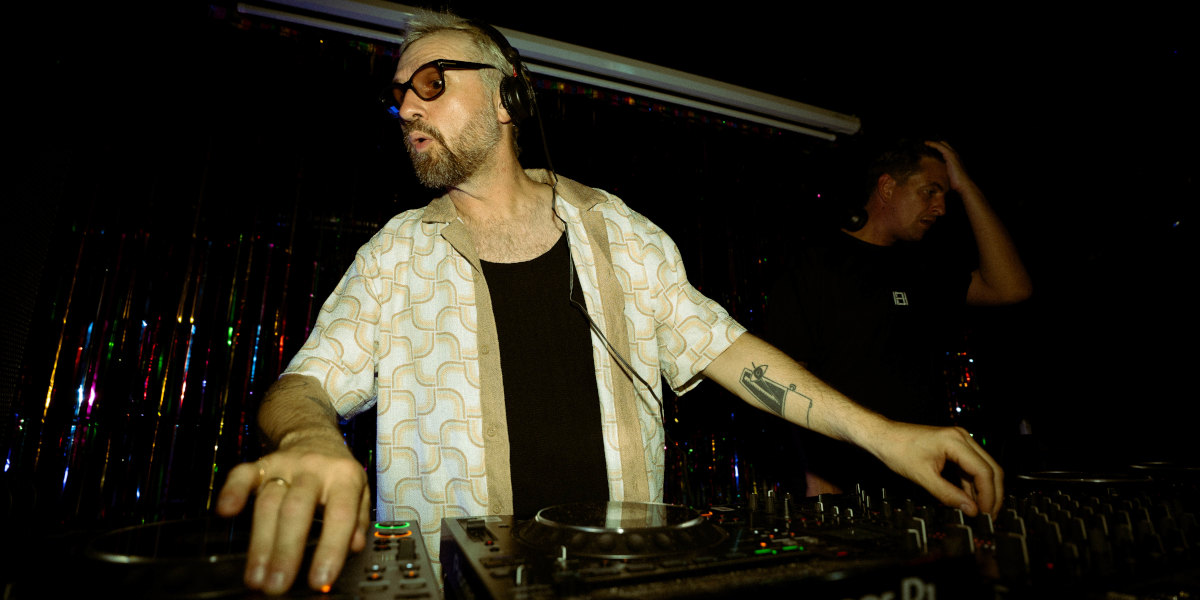 A white man in sunglasses and headphones, wearing a white shirt over black tee, plays the decks against a dark backdrop