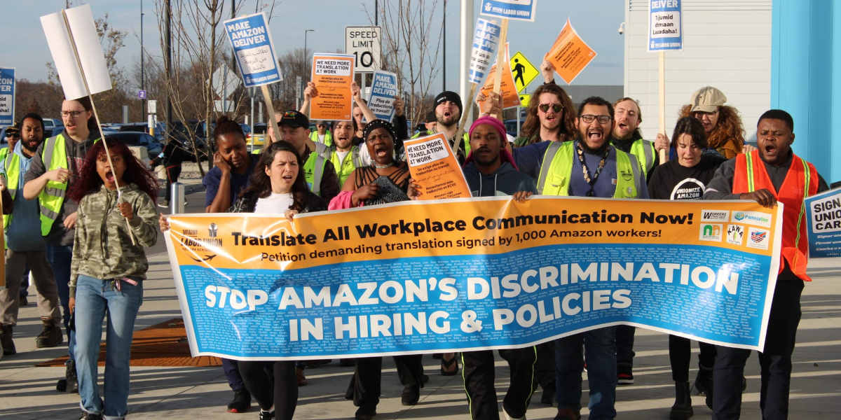 Amazon workers marching behind a banner reading 'Translate all workplace communications now! and 'Stop Amazon's discrimination in hiring & policies'