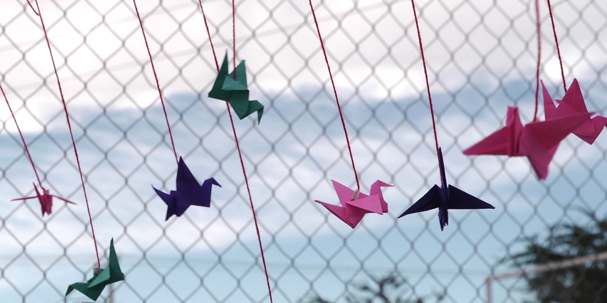 Brightly coloured paper (origami) birds are tied to a chain link fence, with a cloudy blue sky in the background