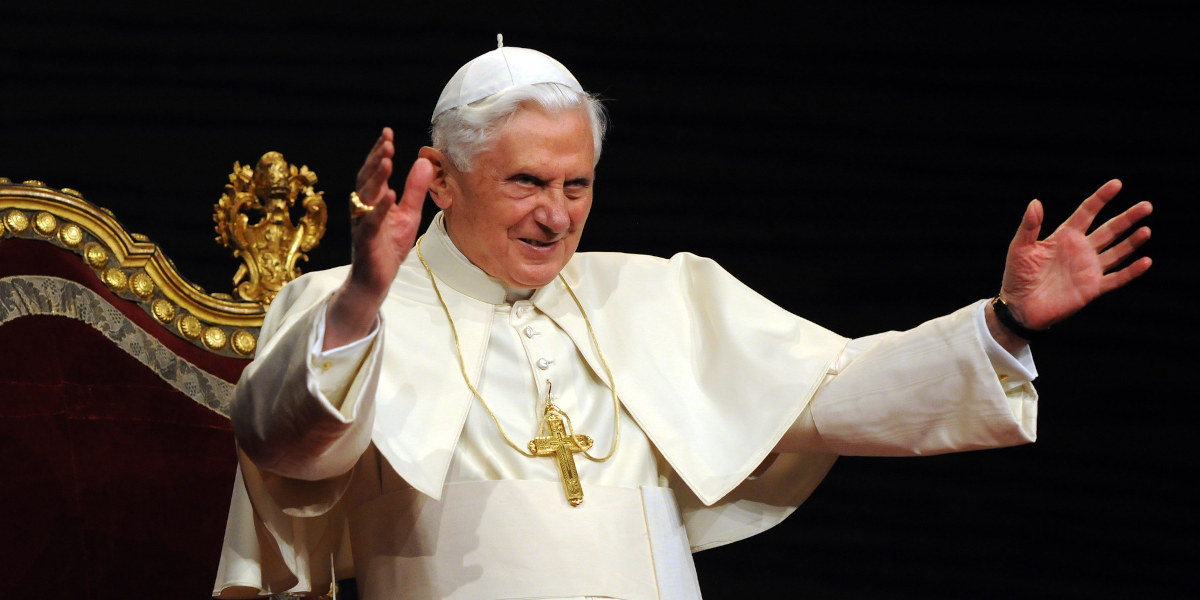 The Pope, dressed in all white robes with a large gold cross and small white hat – raises his arms and smiles to an out of shot audience