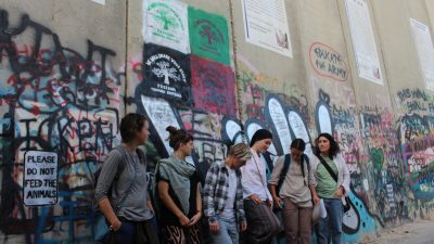 A group of people stand with their backs to a large wall covered in pro-Palestine graffiti and posters