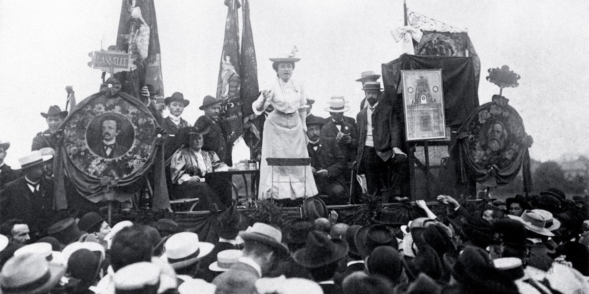 A black and white photograph of Rosa Luxemburg delivering a speech to a crowd