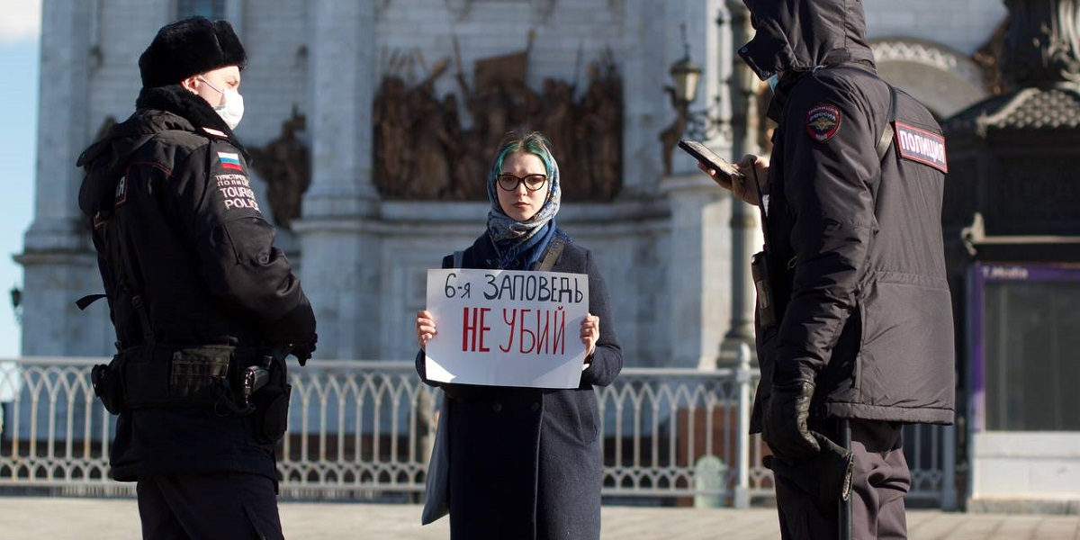 Young woman holds placard that reads 'The Sixth Commandment: Don't Kill', beside Russian police officers