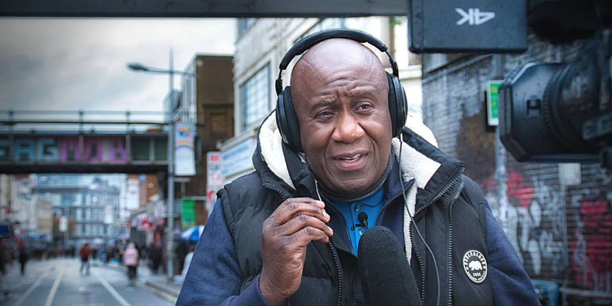 A black man wearing a headset and mic is delivering a report to camera from a city street