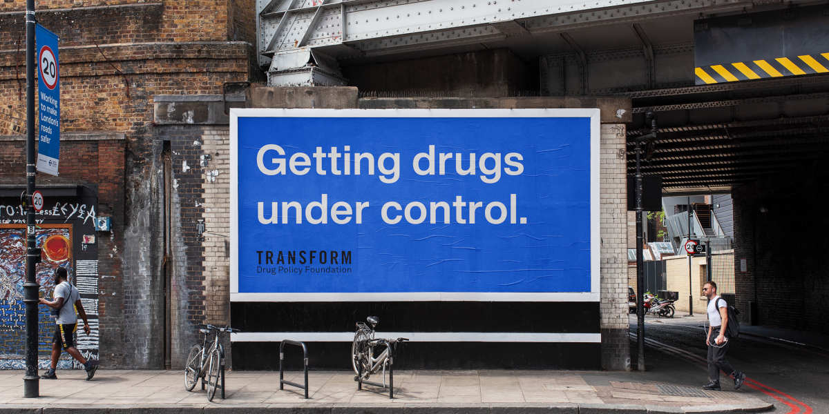 A blue Transform Drug Policy Foundation billboard under a train bridge reading 'Getting Drugs Under Control.'