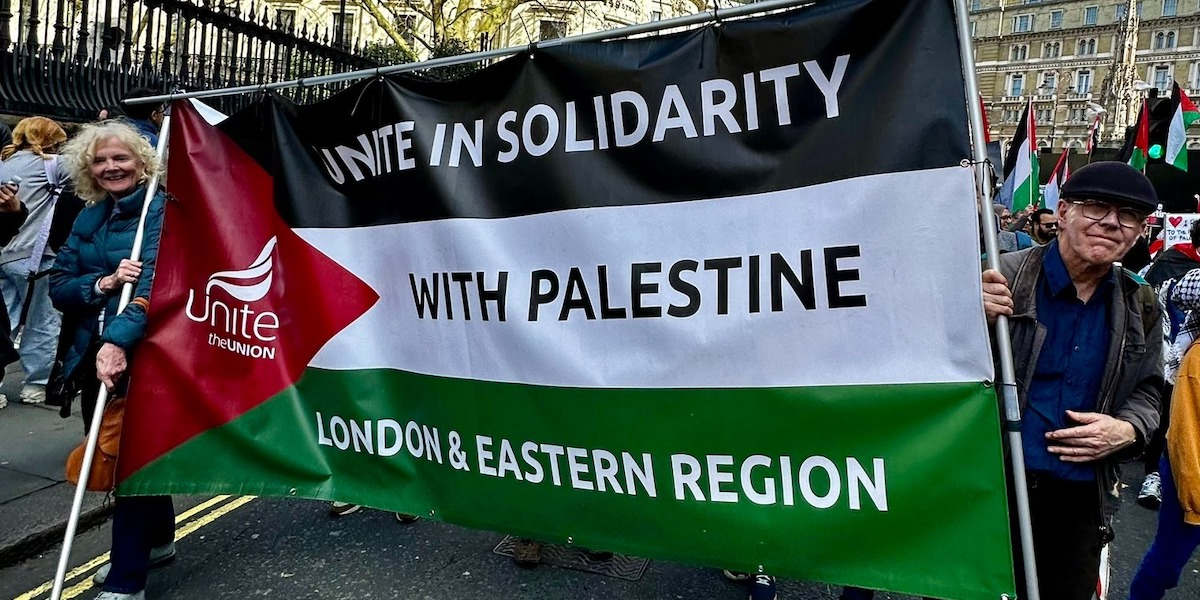 Two members of the Unite union holding a banner bearing the colours of the Palestinian flag an dreading 'in solidarity with Palestine London & Eastern region'