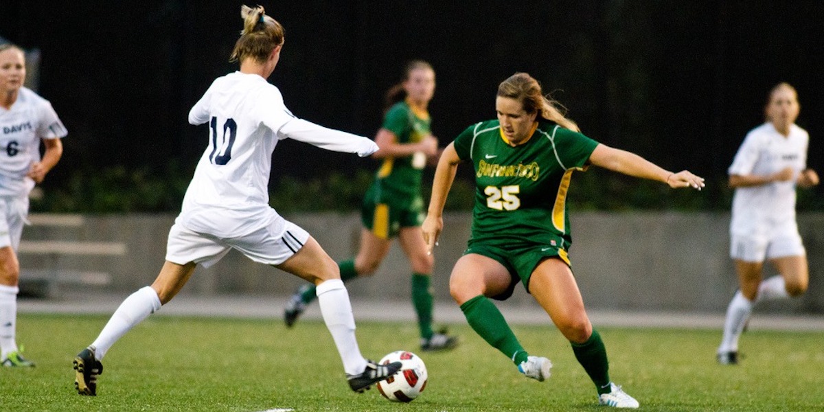 Two women – one in white kit, the other in green – players compete for a football on a pitch during a game
