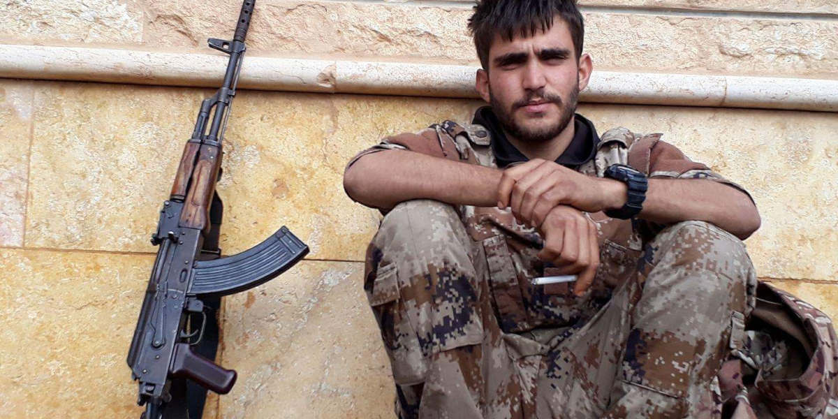 A YPG fighter sitting against a wall resting with his rifle propped up to his left