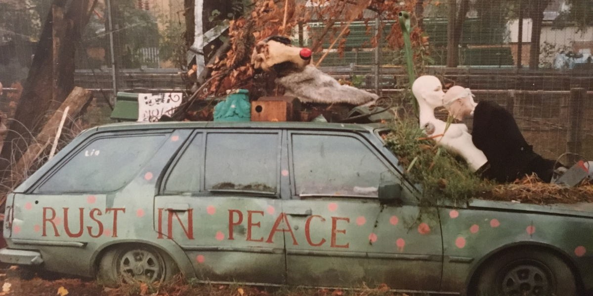 A photograph of an abandoned car with the words 'rust in piece' painted on its side. Two mannequin torsos are resting on the car's bonnet