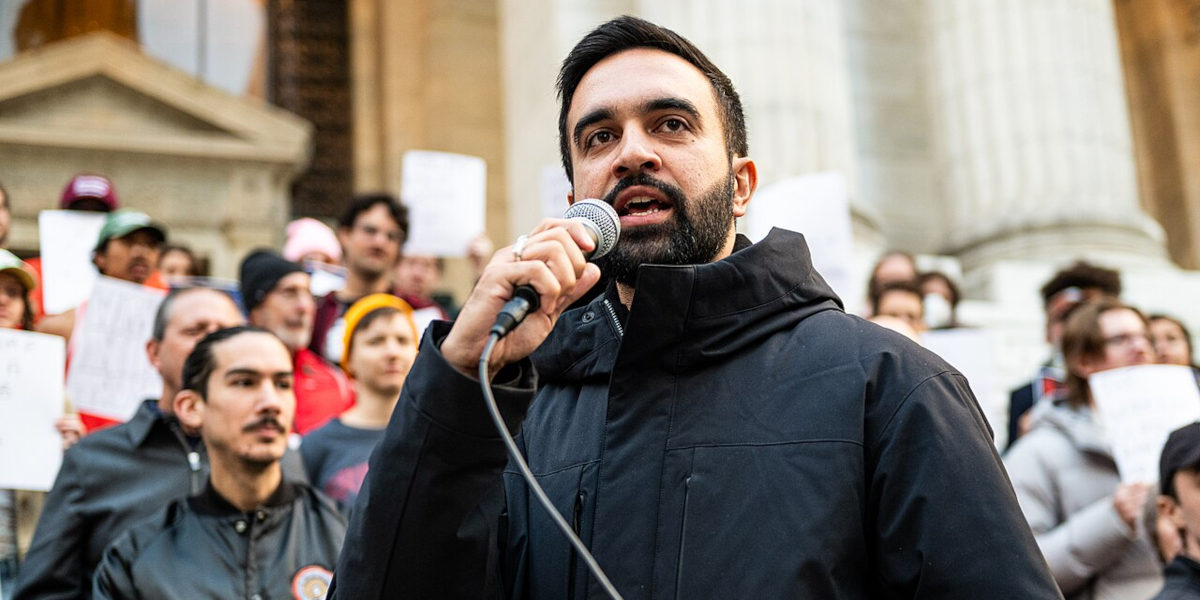Democratic Primary nominee Zohran Mamdani speaking at a Resist fascism rally, with a crowd of people holding signs behind him